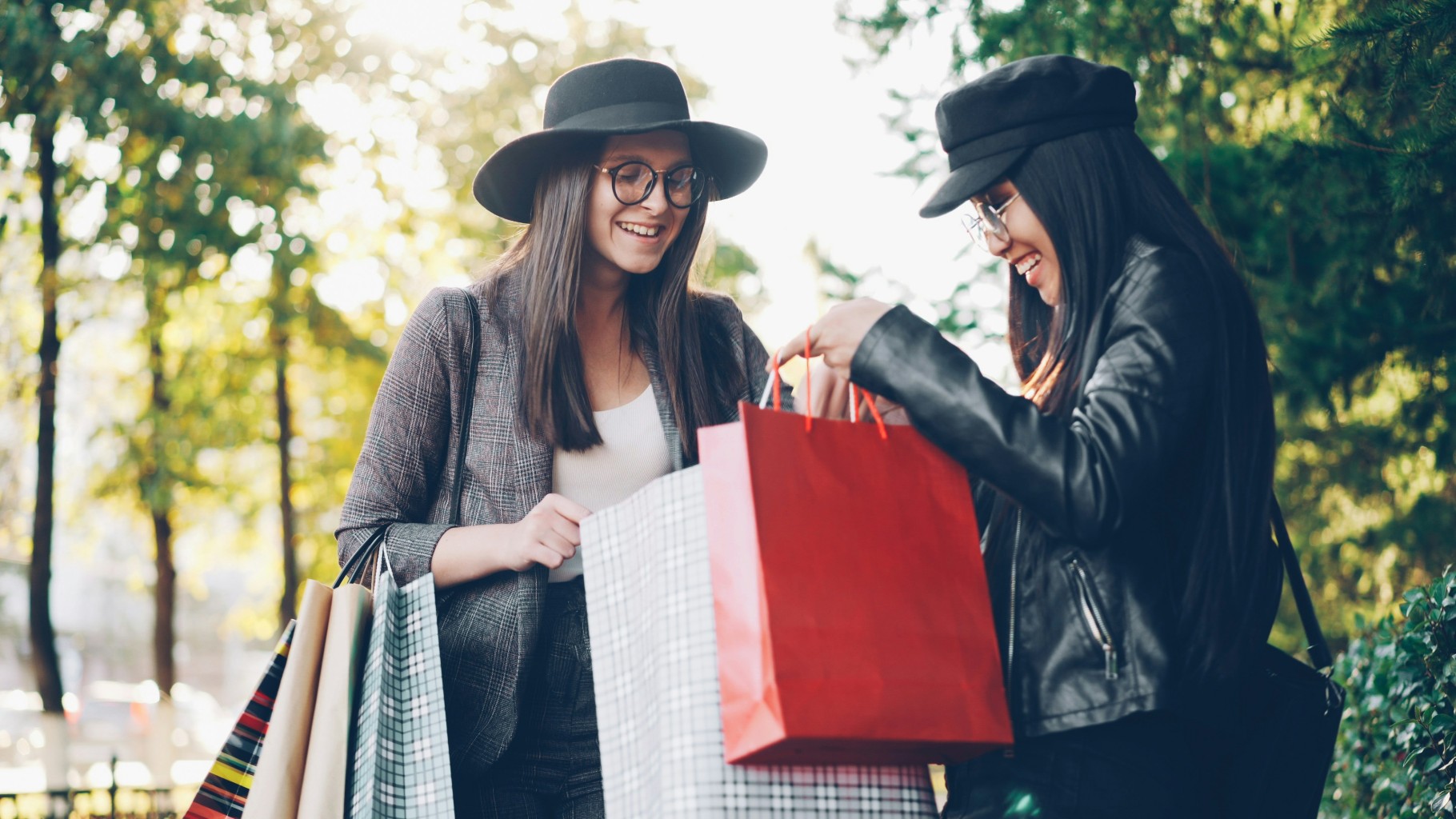 Two gen-z women with shopping bags in a tree-laden street.