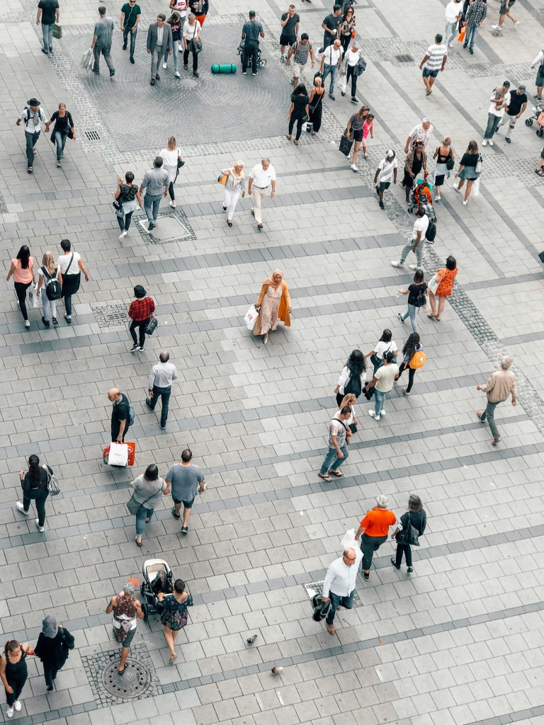 Crowd of people walking through pedestrianised area with shopping bags, prams, backpacks.